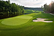 Lush green fairways and sand bunker at Ballantrae Golf Club, bordered by serene woods and a reflective pond under a bright sky.