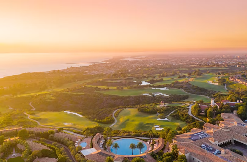 Aerial view of a sunrise over a lush Southern California golf course and oceanfront villas with palm-lined streets.