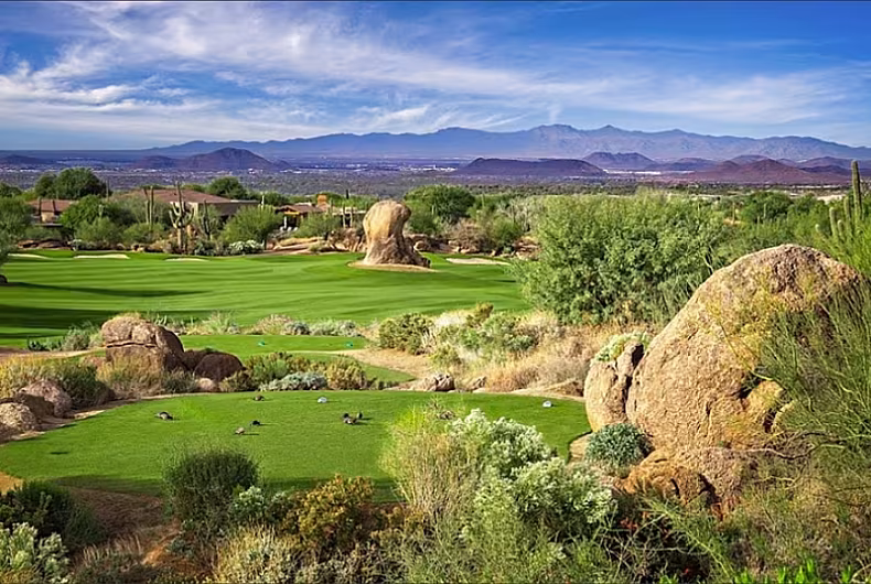 Scenic view of a lush golf course in Scottsdale, surrounded by desert landscape, boulders, and distant mountains under a clear sky.