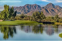 Serene golf course landscape with green fairway, calm water, and distant rugged mountains under a clear blue sky.