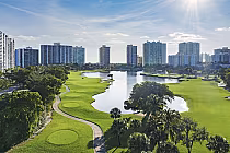 Scenic golf course with lush greens, palm trees, and a pond, surrounded by towering city buildings under a sunny sky.