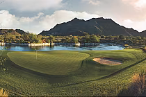 Scenic view of a lush golf course with a sand trap, surrounded by a tranquil lake and mountain backdrop at sunset.