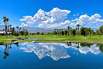 Heritage Palms golf course with lake, palm trees, lush greenery, and mountains under a blue sky with white clouds in Indio.