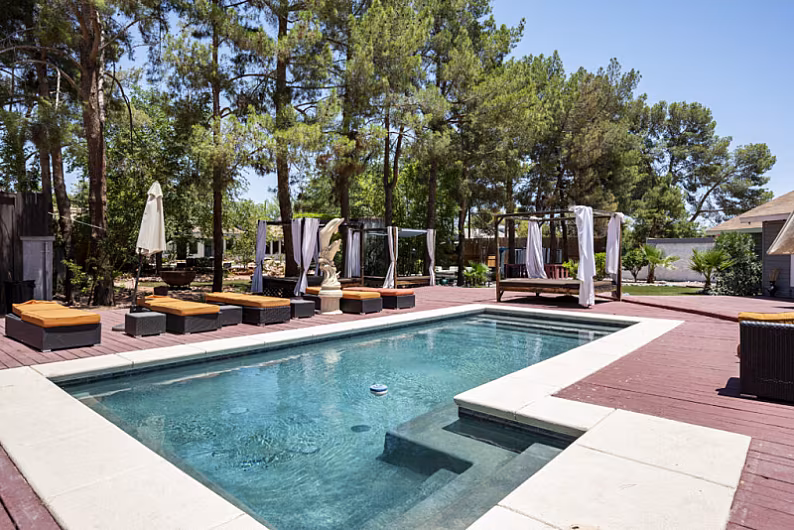 Luxury rental home poolside with cabanas, sun loungers, and surrounding trees under a clear blue Las Vegas sky.