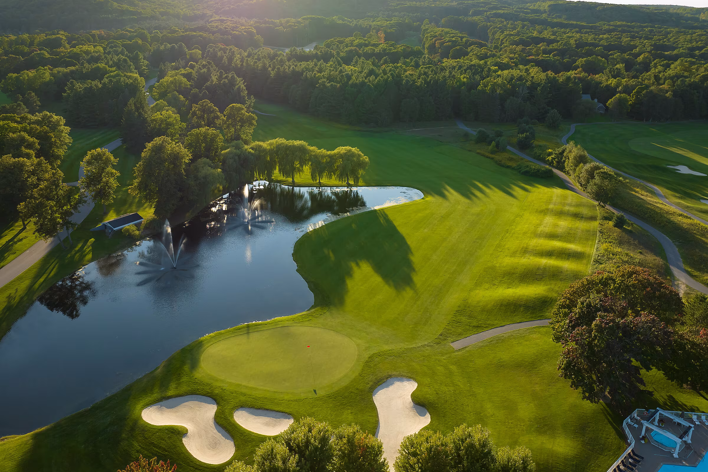 Aerial view of a scenic Michigan golf course featuring lush fairways, a pond with fountains, and surrounded by dense forest.