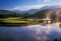 Scenic view of Nicklaus North Golf Club with lush greens, a stone bridge, and mist over a tranquil lake, surrounded by mountains.