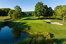 Lush green golf course at Boyne Mountain, featuring sand bunkers, a pristine pond, and surrounding trees under a clear sky.