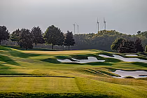 Lush green golf course at The Hawk Golf Club, featuring sand bunkers, manicured lawns, and trees with wind turbines in the background.