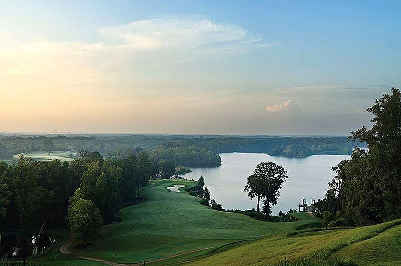 Scenic view of a golf course with lush green fairways, trees, and a large body of water under a partly cloudy sky.