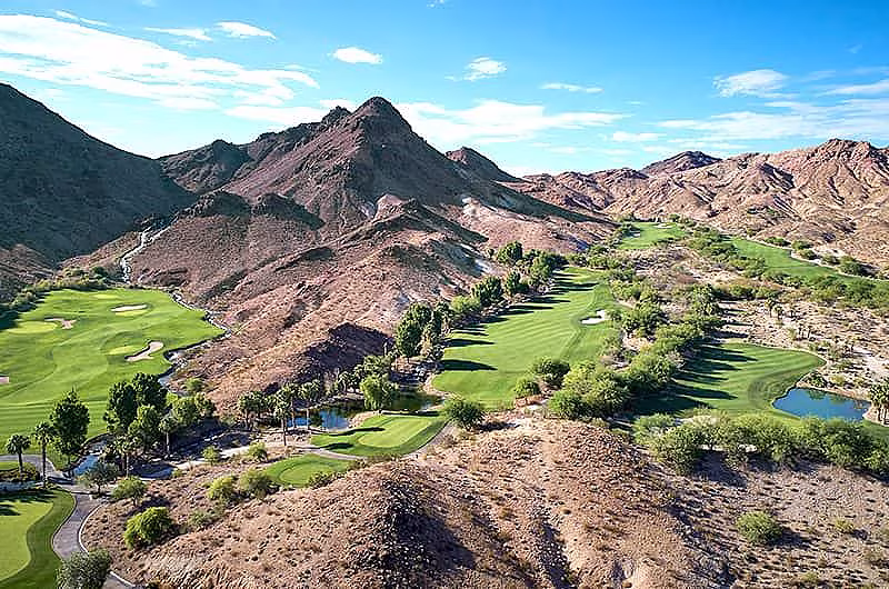A scenic view of a golf course with lush green fairways and water hazards, set against rugged desert mountains under a blue sky.