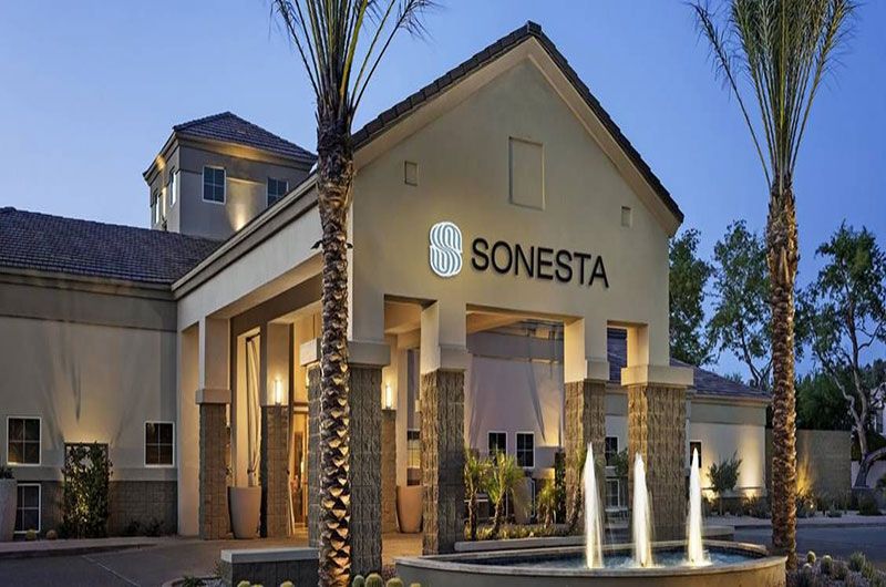 Sonesta Suites Scottsdale hotel exterior at dusk with illuminated sign, palm trees, and water fountain in front.