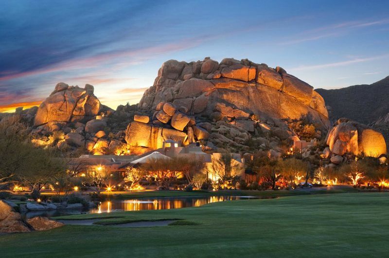 The Boulders Resort and Spa at sunset, featuring illuminated large rocks, lush green golf course, and a serene water feature.