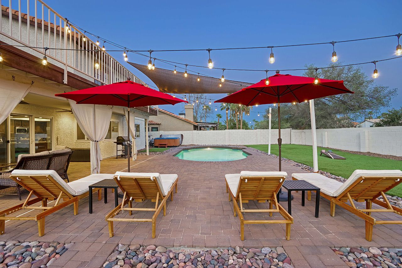 Luxury Scottsdale vacation home backyard with a heated pool, hot tub, sun loungers, umbrellas, string lights, and a grassy area.