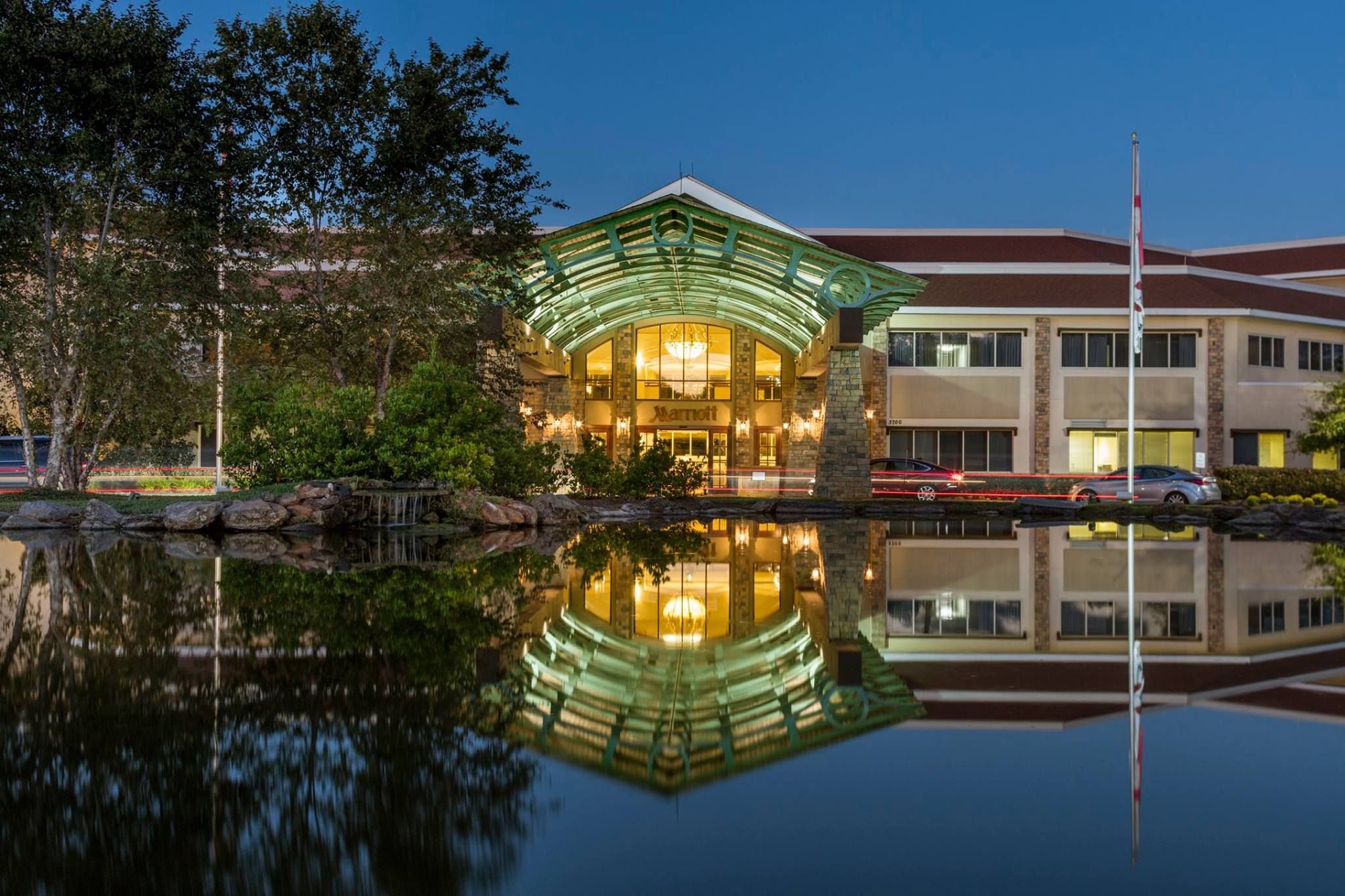 Night view of Auburn Marriott Opelika Resort with illuminated entrance, reflected in a calm pond, surrounded by trees and cars.