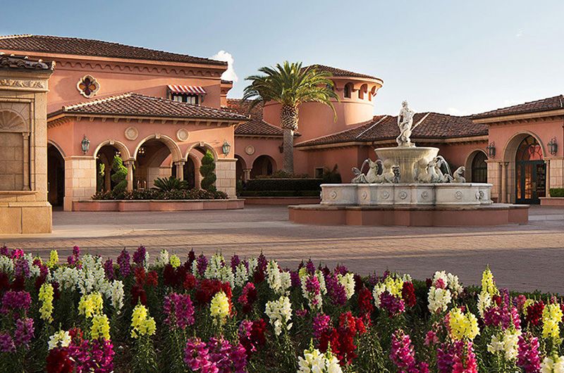 Lavish pink Mediterranean-style building with a fountain, palm trees, and colorful flowers at Fairmont Grand Del Mar golf retreat.