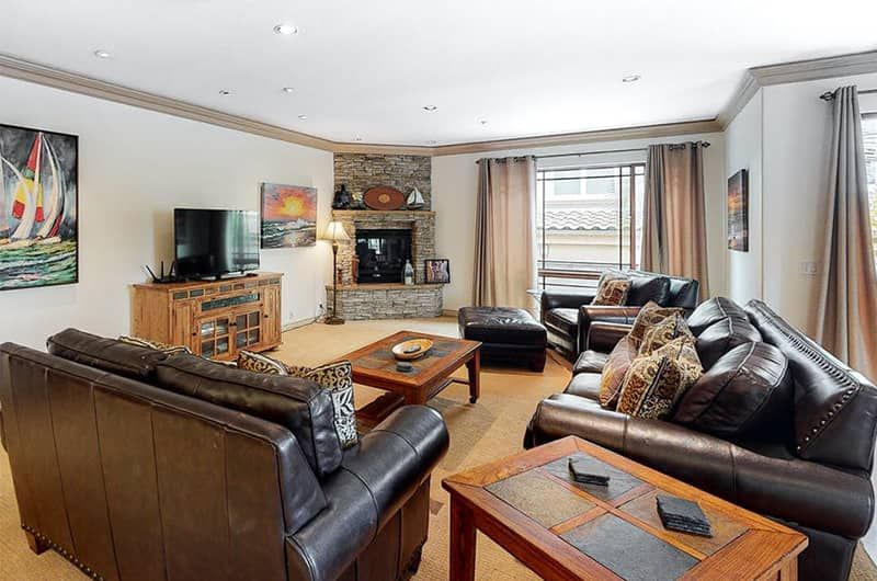 Spacious living room with leather sofas, a stone fireplace, large windows with curtains, and a flat-screen TV, in a luxurious vacation home.