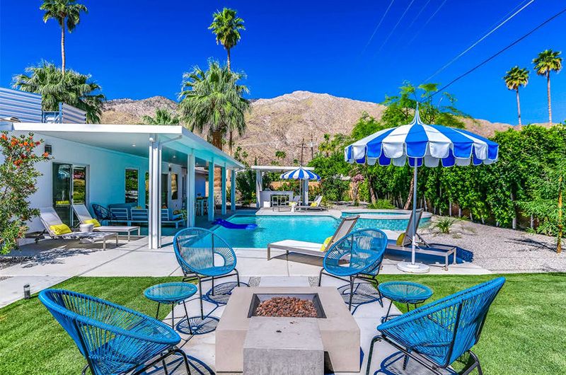 Desert Stardust Luxurious desert home with a private pool, outdoor firepit, lush greenery, palm trees, and mountain backdrop under a clear blue sky.