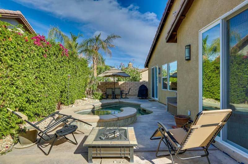 Indian Palms Vacation Villa Outdoor patio of a vacation villa with a hot tub, lounge chairs, a table with an umbrella, lush greenery, and large glass doors.