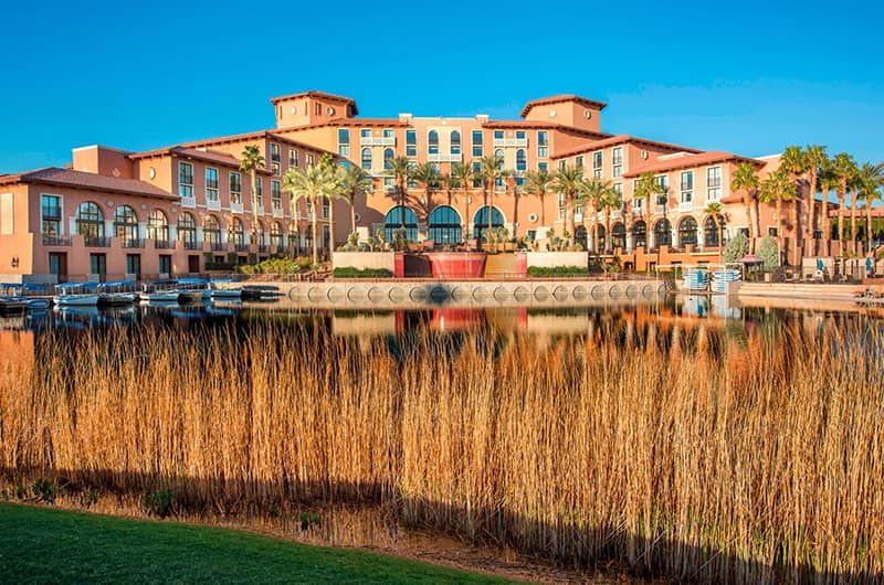 Westin Lake Las Vegas Luxury resort with pink stucco buildings, palm trees, a water fountain, and boats on a lake under a clear blue sky.