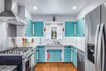 Oliver's Oasis Bright kitchen with turquoise cabinets, white walls, stainless steel appliances, and a window with floral curtains.