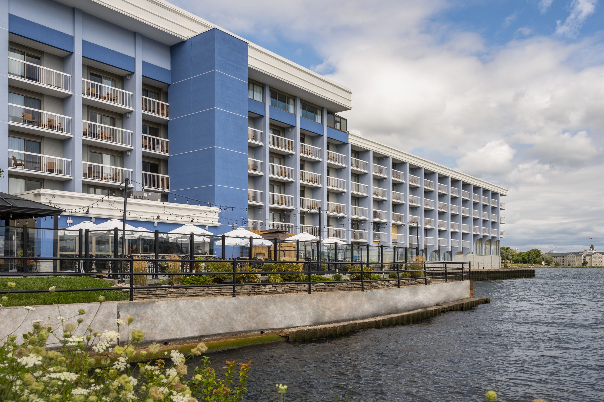 Holiday Inn Waterfront Kingston Luxury hotel by the water with balconies, outdoor seating, and string lights, overlooking a river under a partly cloudy sky.