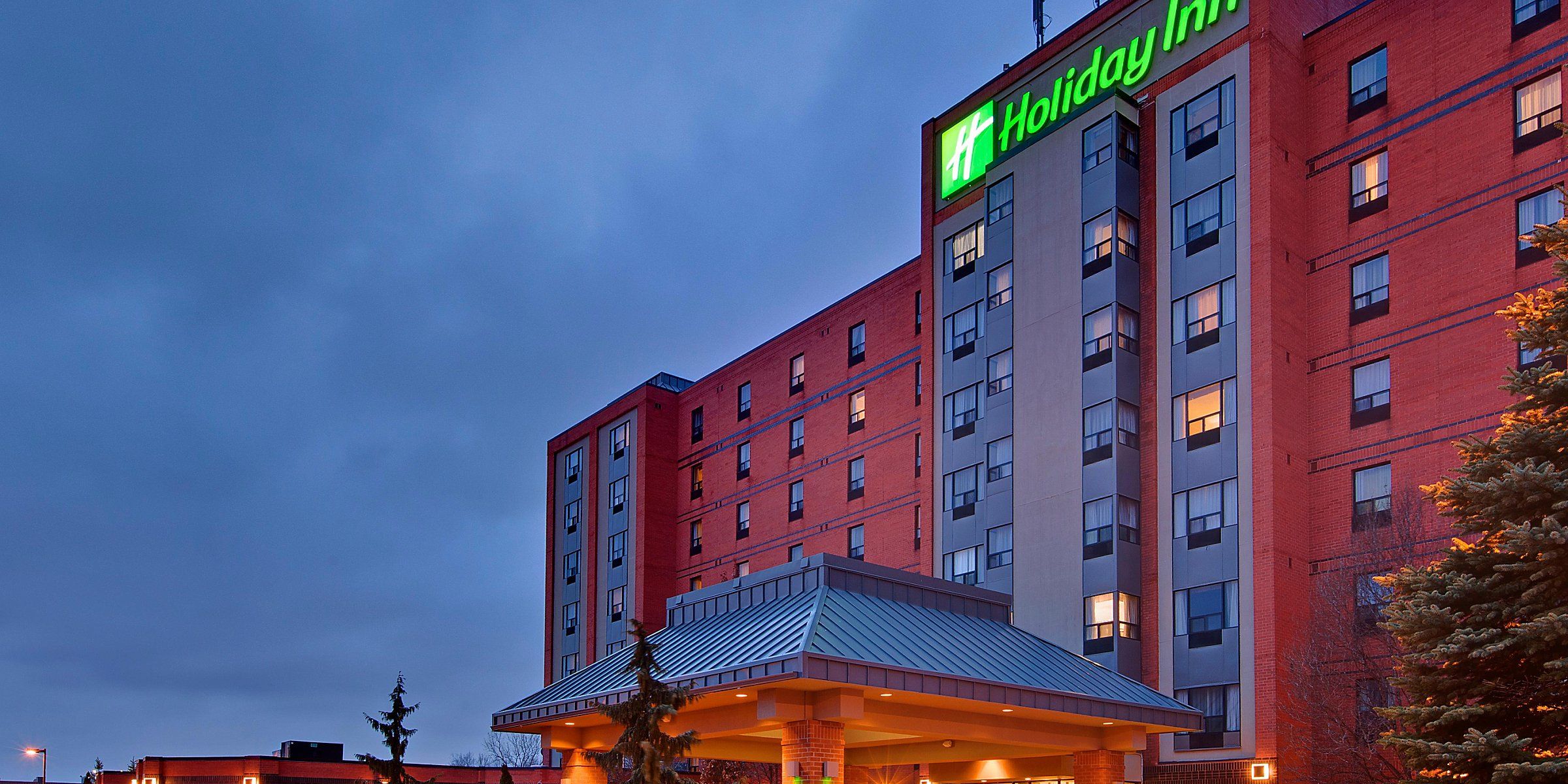 Holiday Inn Ambassador Bridge Night view of the Holiday Inn hotel with illuminated green sign, red brick exterior, and lit entrance, near Windsor golf courses.