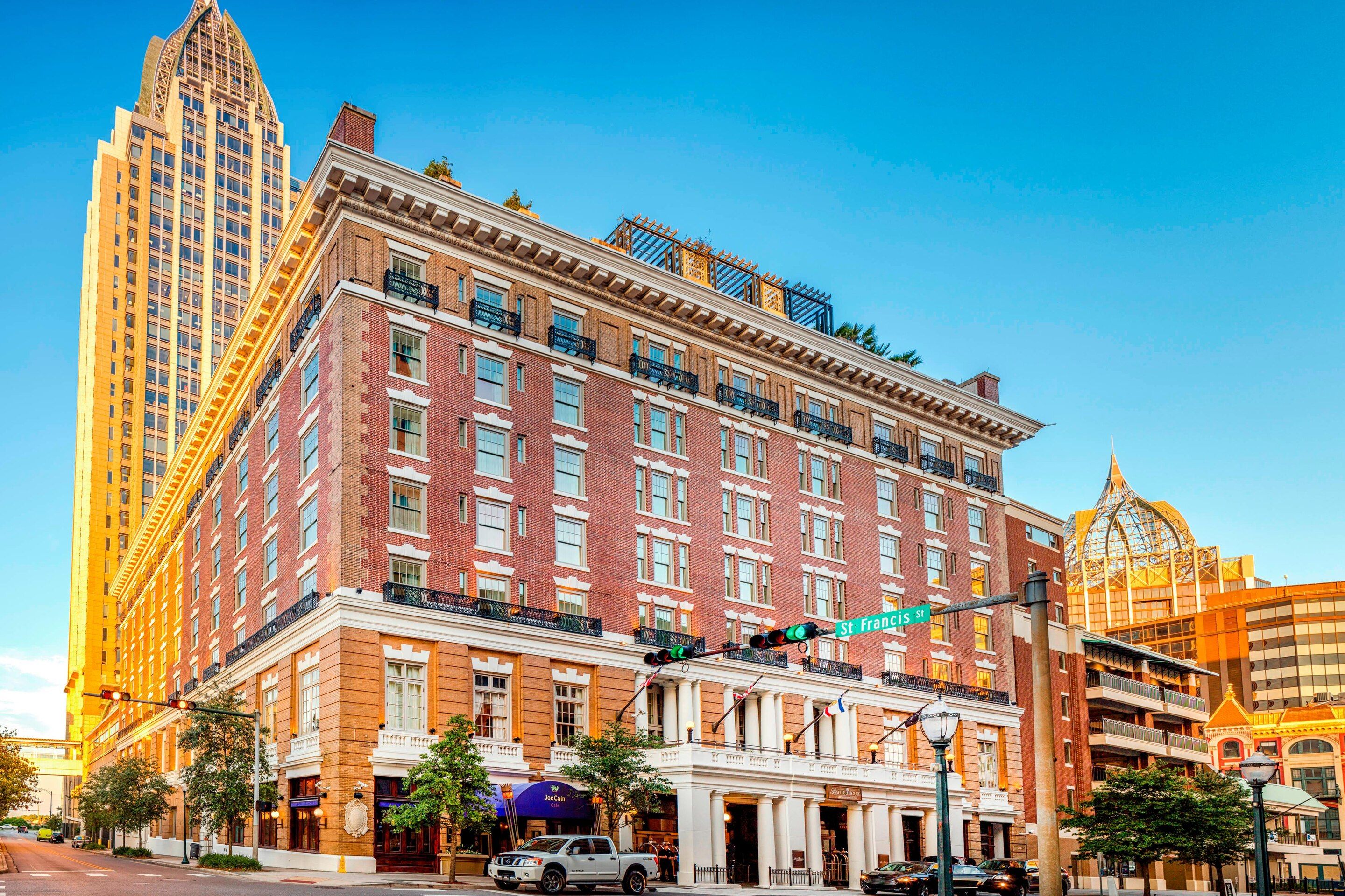 Historic hotel building with brick facade, white columns, and balconies, located on a city street with tall skyscrapers nearby.