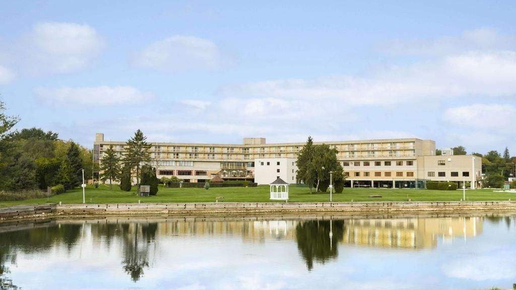 Ramada by Wyndham Belleville Hotel building with multiple floors, surrounded by green lawns, trees, and a water body reflecting the structure, under a partly cloudy sky.