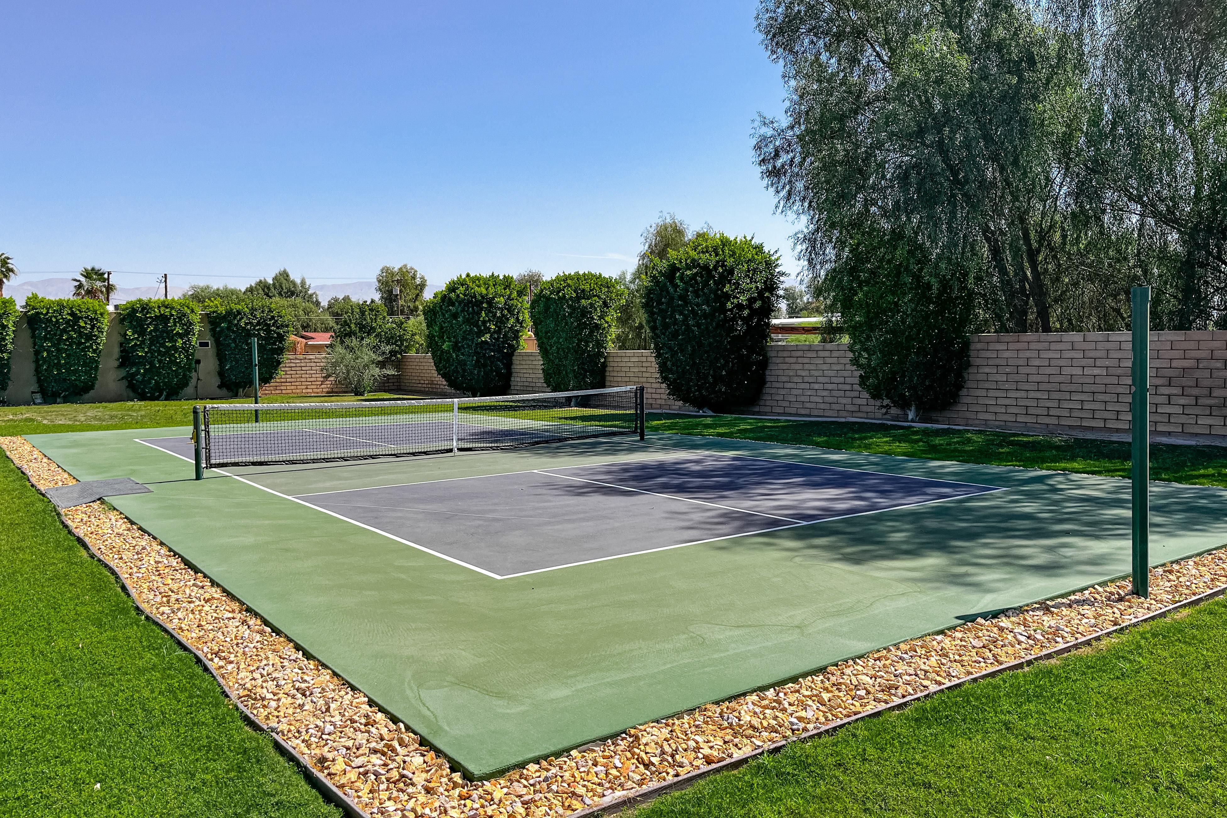 Pickleball Ranch A well-maintained pickleball court with green and gray surface, surrounded by lush trees and a brick wall, sunny sky.