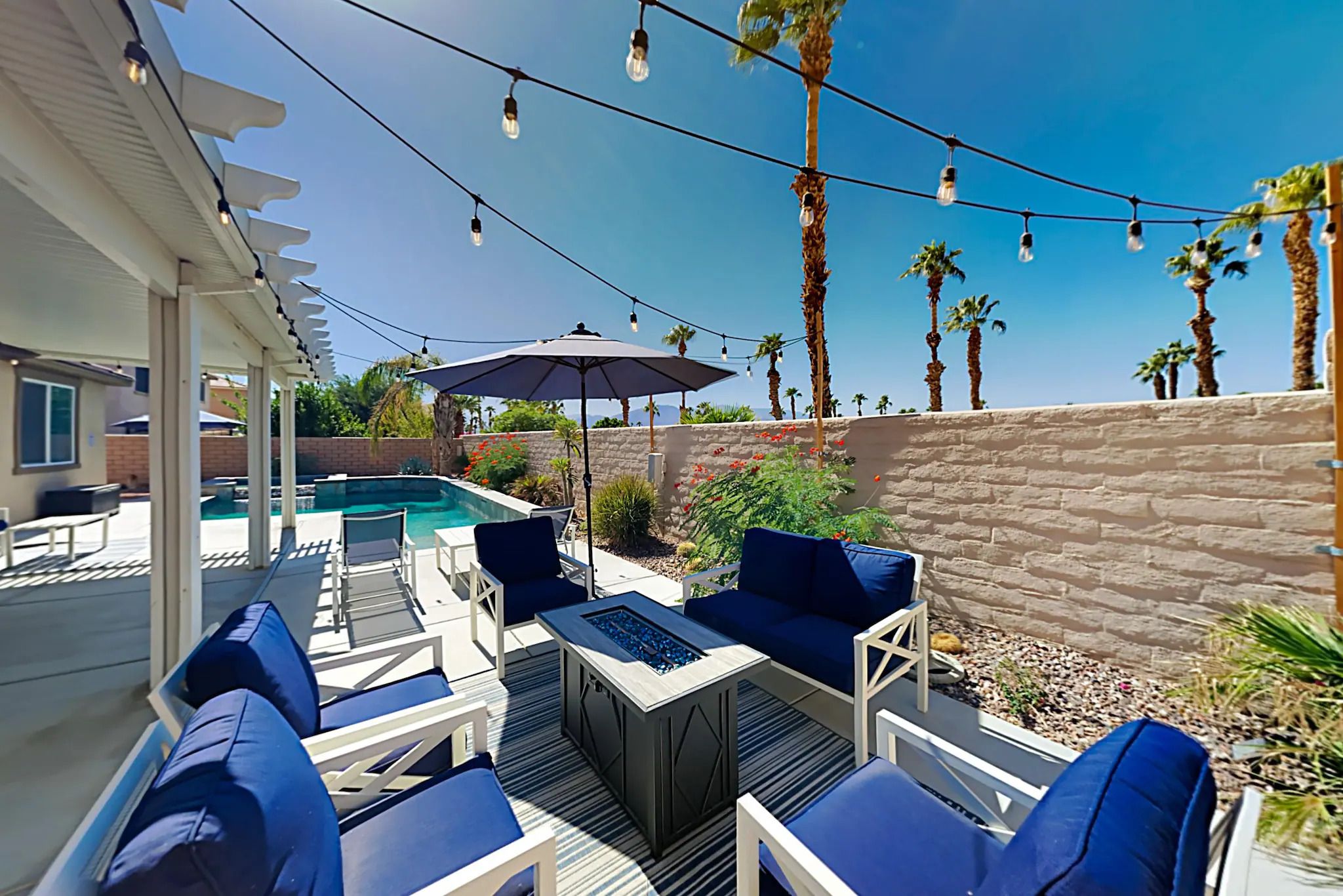 FIORE HAVEN Luxurious backyard with a swimming pool, outdoor seating, umbrella, string lights, palm trees, and a clear blue sky.