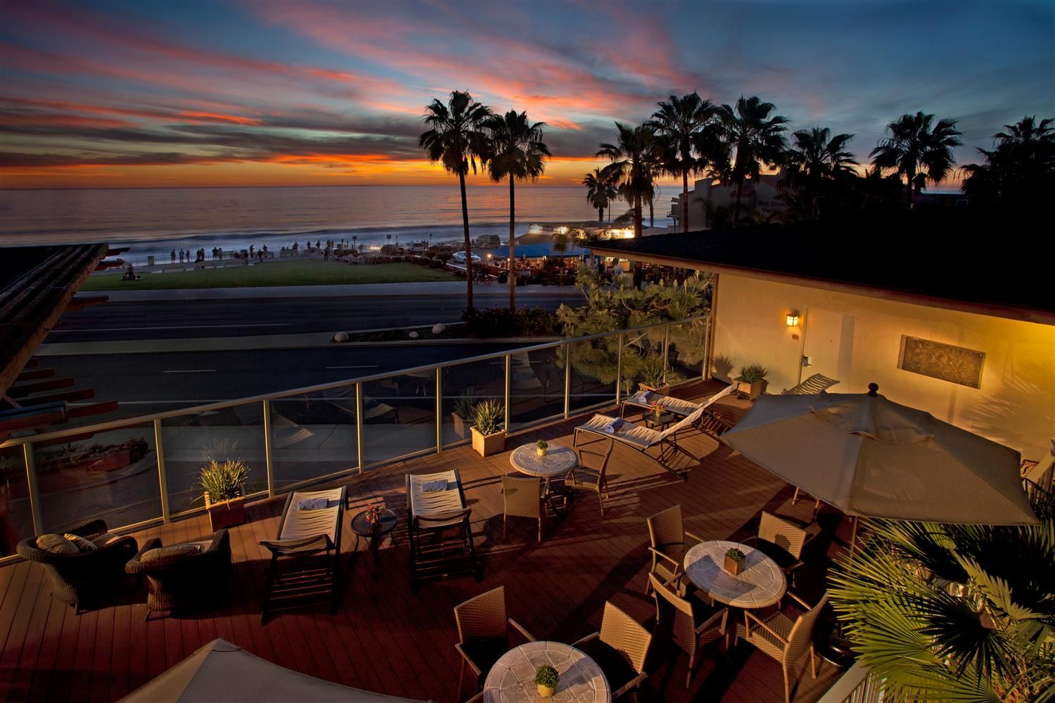 A scenic view from a hotel balcony overlooking Carlsbad Beach at sunset, with palm trees, outdoor seating, and the ocean in the background.