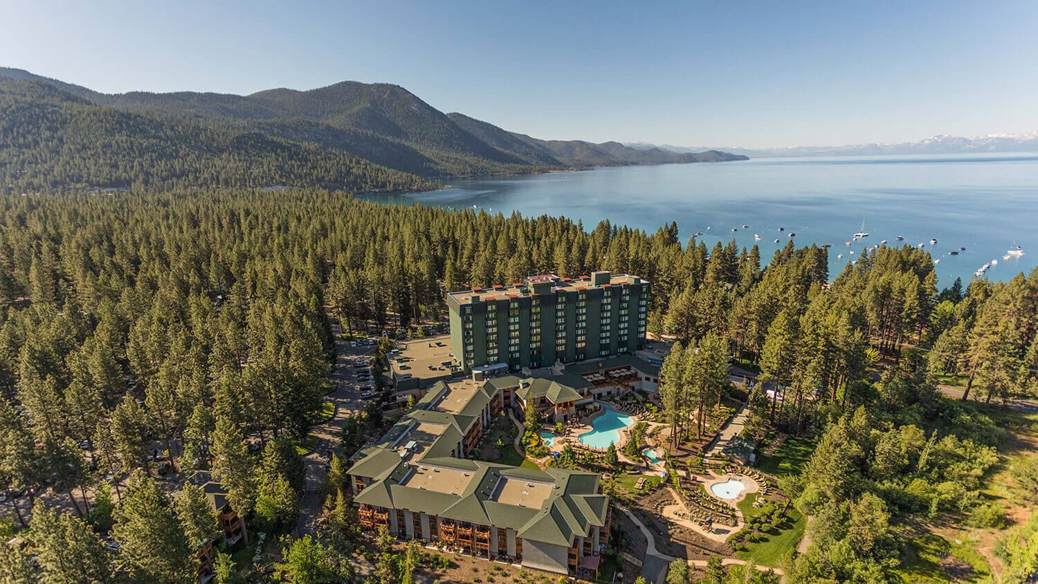 Aerial view of Hyatt Regency Lake Tahoe resort surrounded by pine trees, with a large lake and mountains in the background.