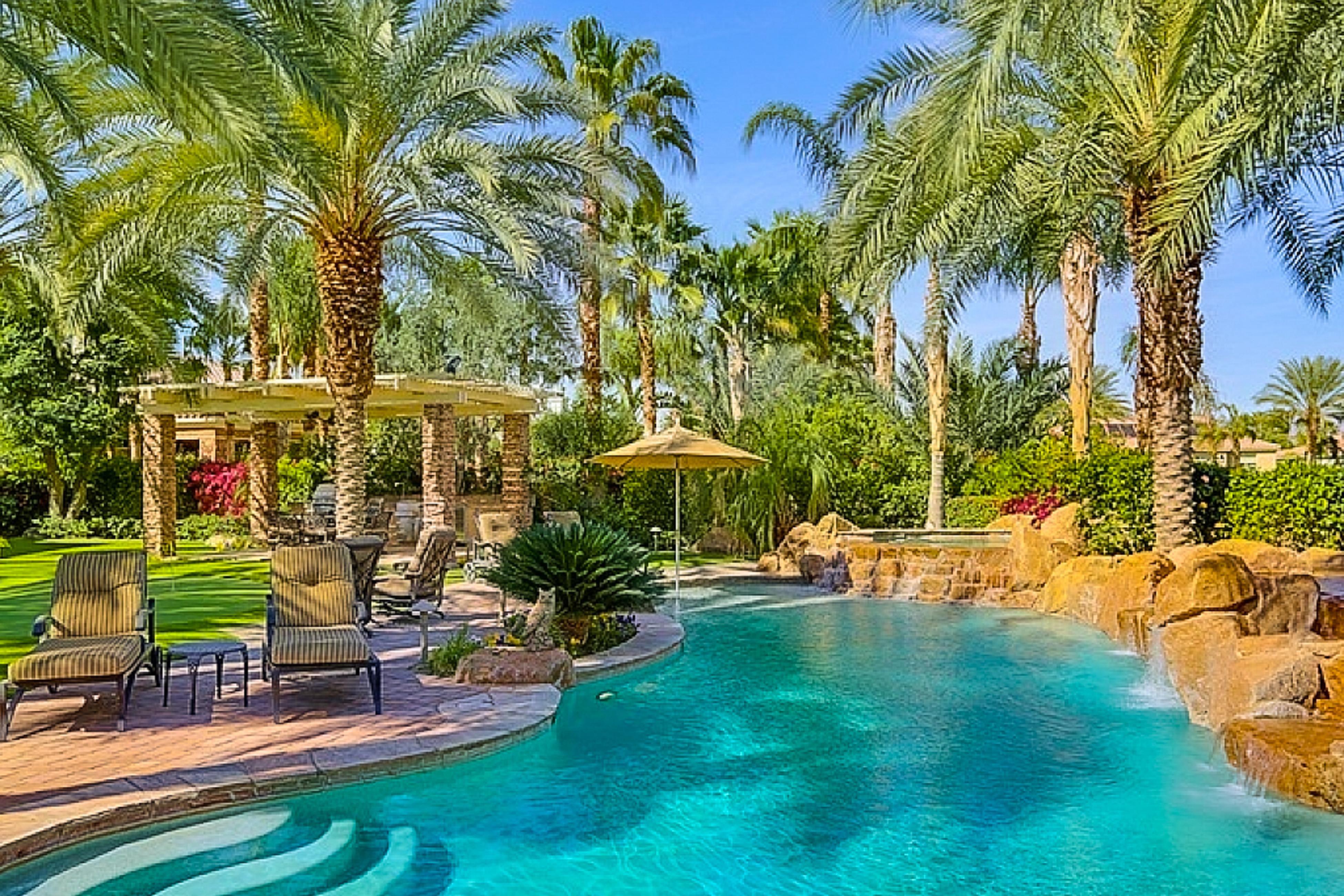 Legends Retreat Luxury backyard pool area with palm trees, lounge chairs, umbrella, and a waterfall feature in a tropical setting.