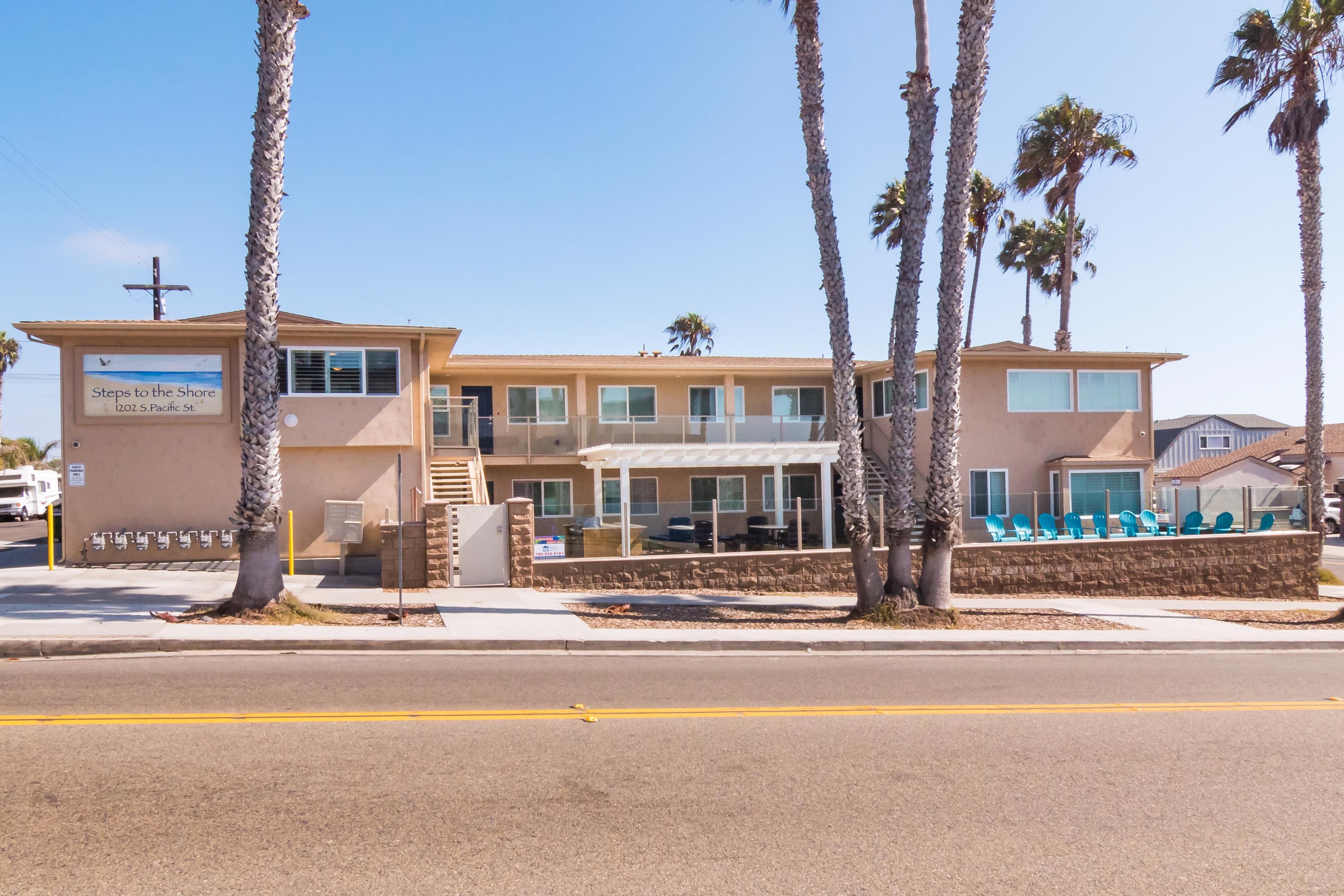 Beachfront condo with large windows, outdoor seating, palm trees, and a clear blue sky in Oceanside, California.