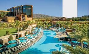 Luxury resort pool area with lounge chairs, palm trees, and a modern hotel building in the background, offering panoramic views.