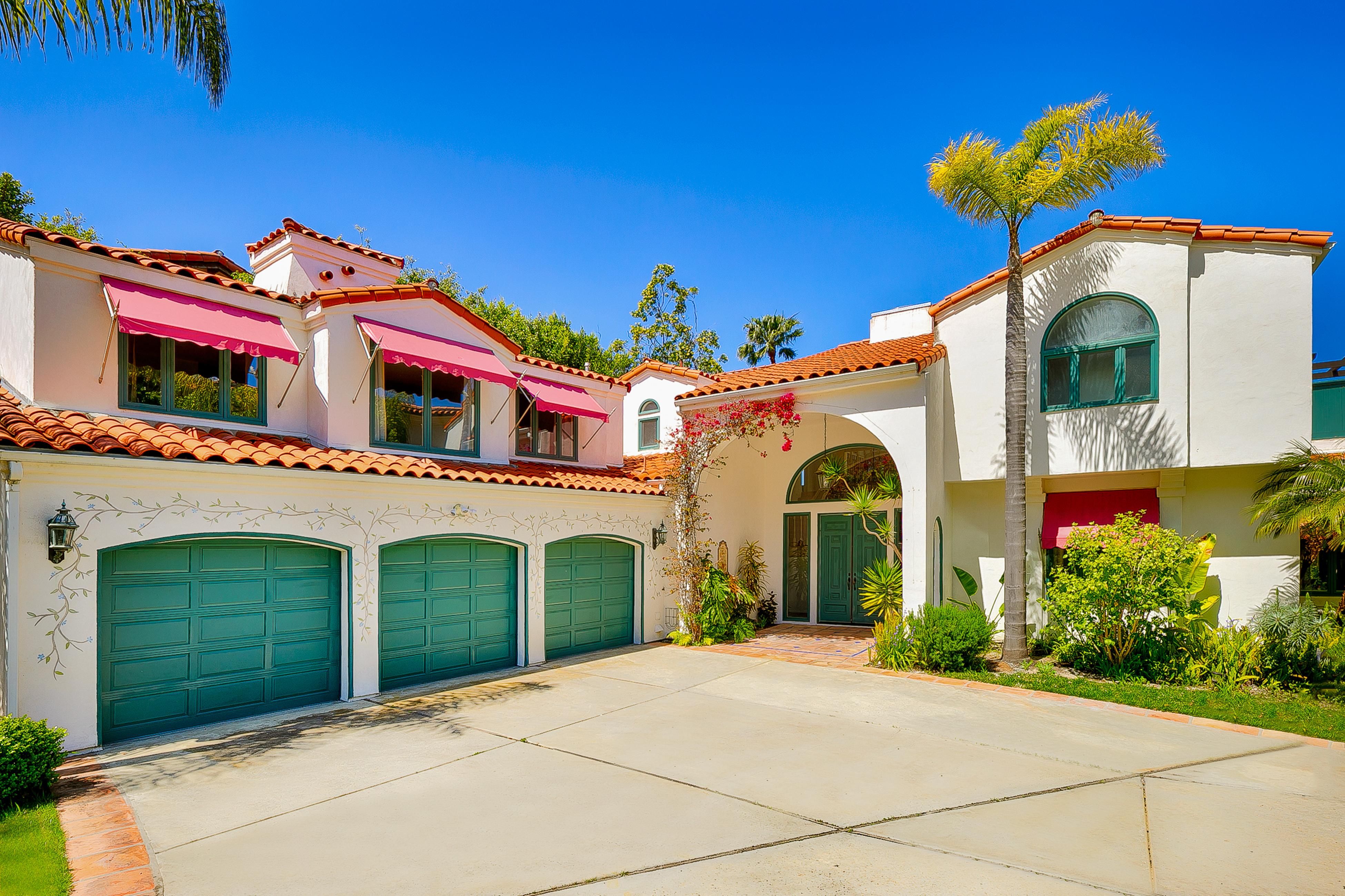 Luxurious 7-bedroom La Jolla home with green garage doors, palm trees, and vibrant landscaping under a clear blue sky.