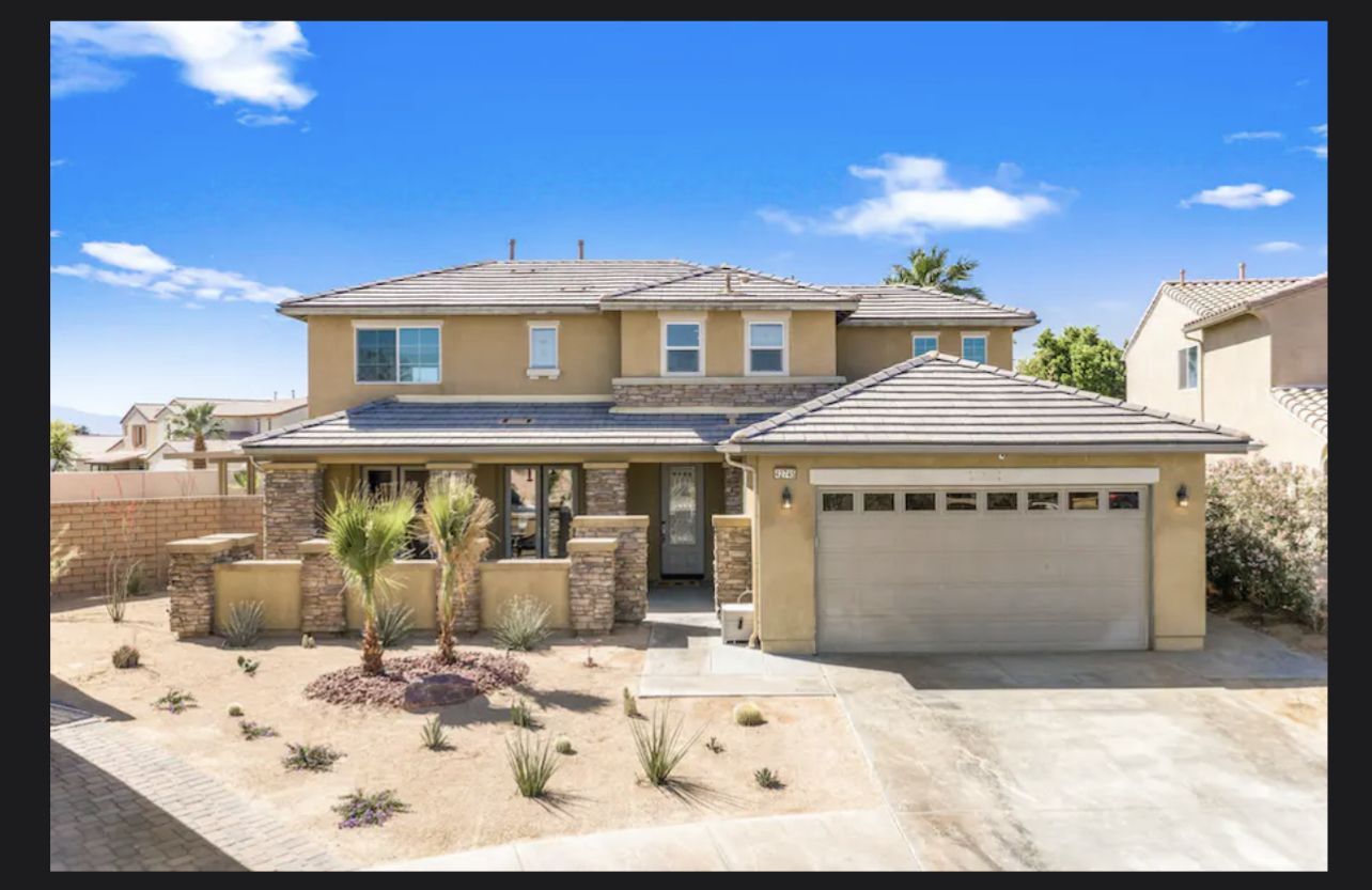 Casa de Luxe - Indio, CA A two-story luxury home in Indio, California, with a beige exterior, tiled roof, a two-car garage, desert landscaping, and a clear blue sky.