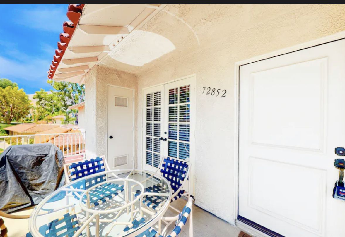 Easy Living Palm Desert - Palm Desert, CA Sunny condo balcony in Palm Desert with white doors, blue and white patio furniture, and a barbecue grill, overlooking lush greenery.