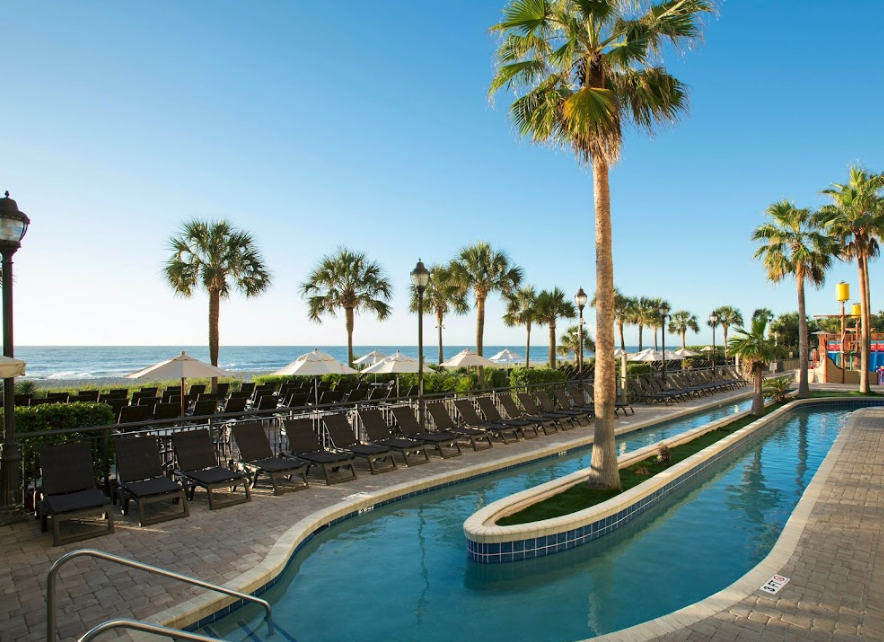 Breakers Resort Poolside view at The Breakers Resort in Myrtle Beach with palm trees, lounge chairs, umbrellas, and ocean in the background.