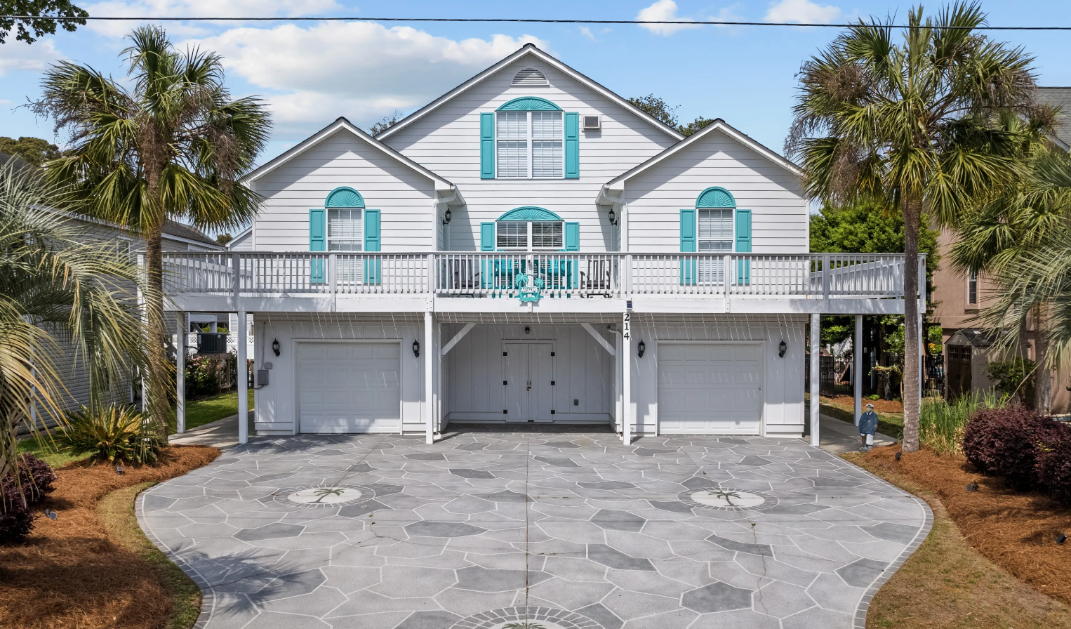 Coastal "N" Coastal luxury vacation home in Surfside Beach, SC with white exterior, blue shutters, large balcony, palm trees, and a patterned driveway.
