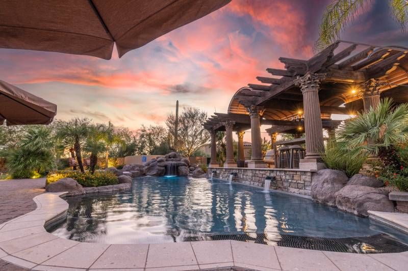 Luxurious Scottsdale vacation home with a pool, waterfall, and classical columns, set against a colorful sunset sky.