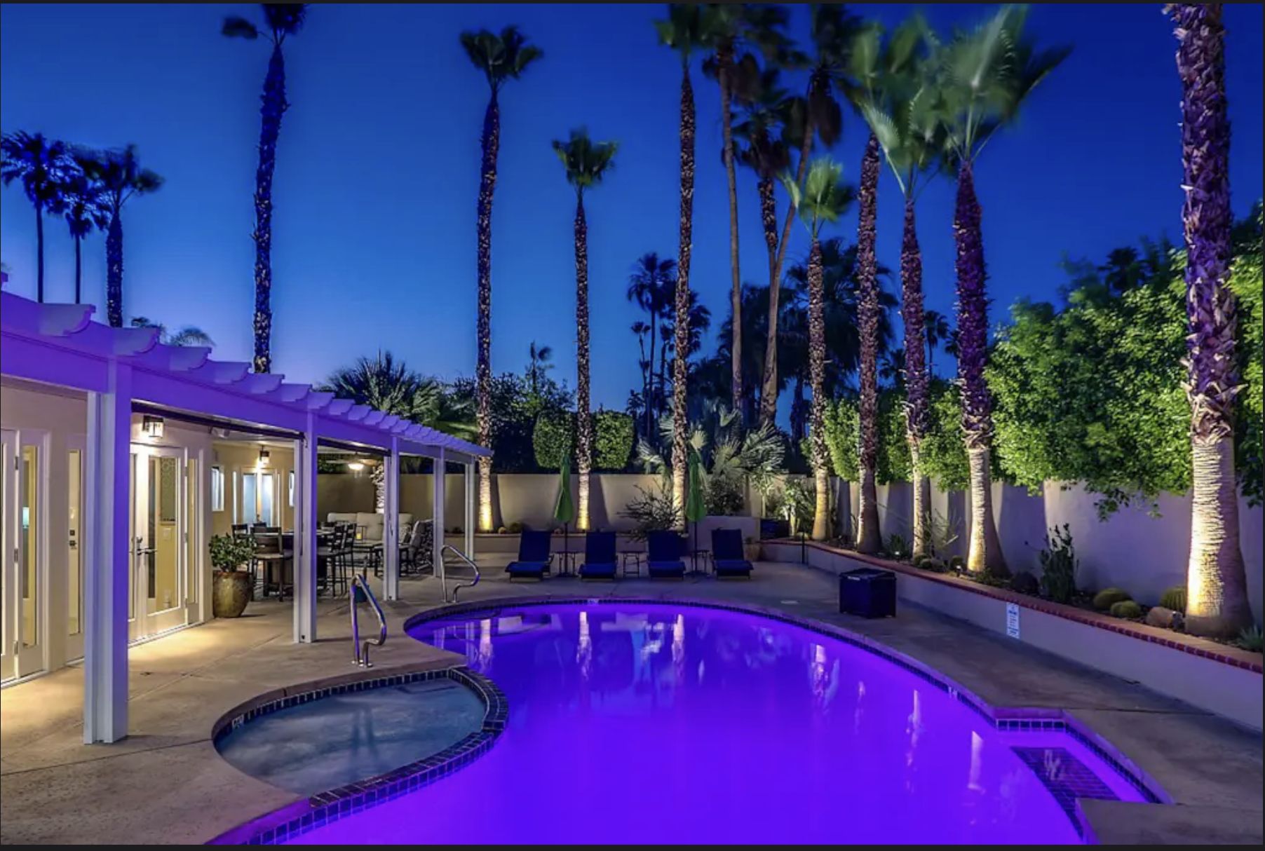 Casa De Myah - Palm Springs, CA Night view of a luxurious backyard with a lit swimming pool, palm trees, outdoor seating, and a private pool and spa at Casa de Myah in Palm Springs.