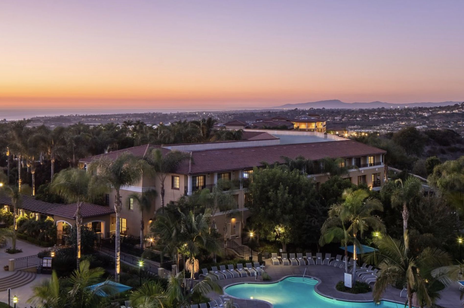 A luxurious coastal resort in Carlsbad, CA, featuring a swimming pool, lush palm trees, and scenic sunset views over the ocean.