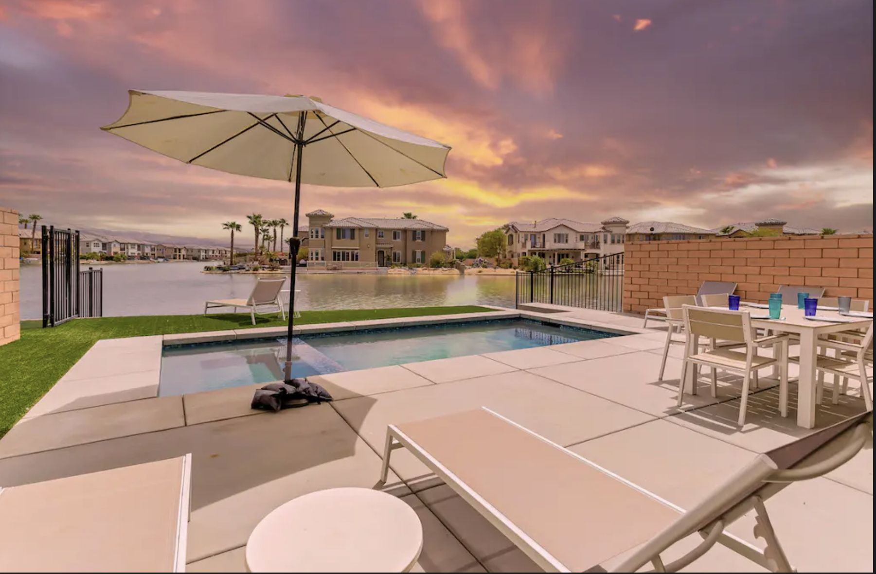 Lakeside Tranquility - Indio, CA Lakeside house in Indio, CA with a pool, outdoor dining area, lounge chairs, and a scenic water view at sunset.