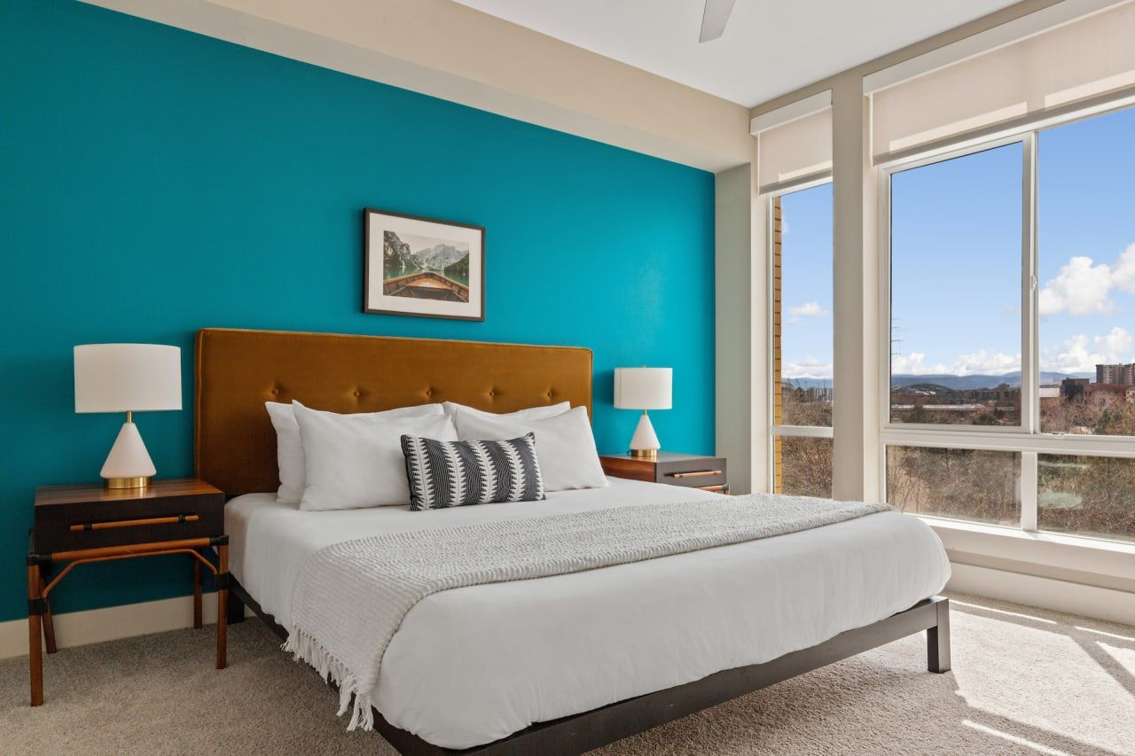 Modern bedroom in Denver's Kasa Union Station apartments with a teal accent wall, large windows, and city views.
