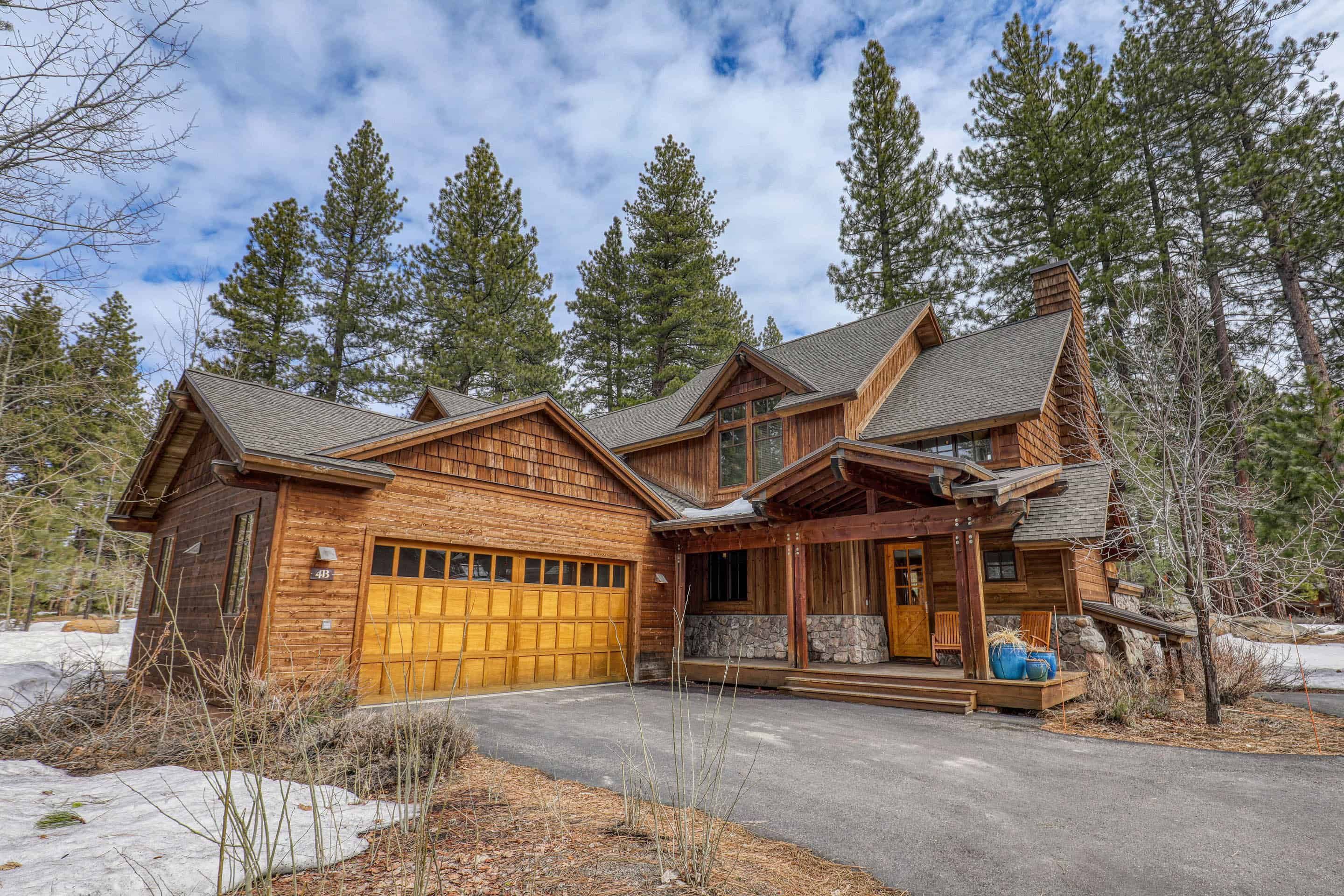 Luxury wooden cottage at Old Greenwood with a garage, surrounded by tall pine trees and a partly cloudy sky.