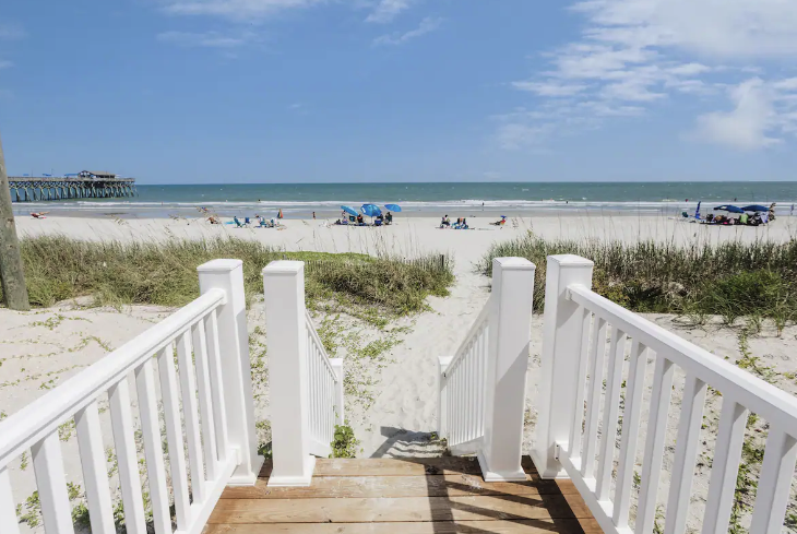 Pirates Cove View from a wooden staircase leading to a sandy beach with beach chairs, umbrellas, and the ocean under a partly cloudy sky.