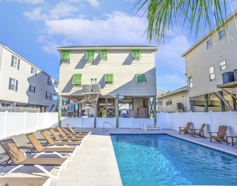 The Salty Dolphin Luxury beach house with a heated pool, surrounded by lounge chairs, near Garden City pier, with colorful window awnings.