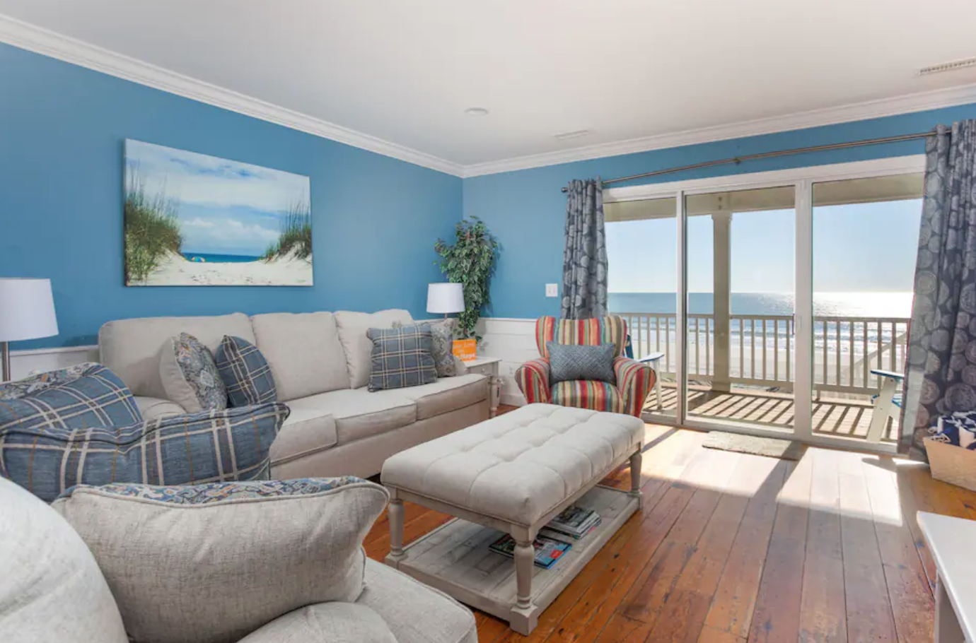 Mermaids Haven Living room with blue walls, beige sofa, colorful armchair, sliding glass doors to balcony with ocean view, and wooden floor.