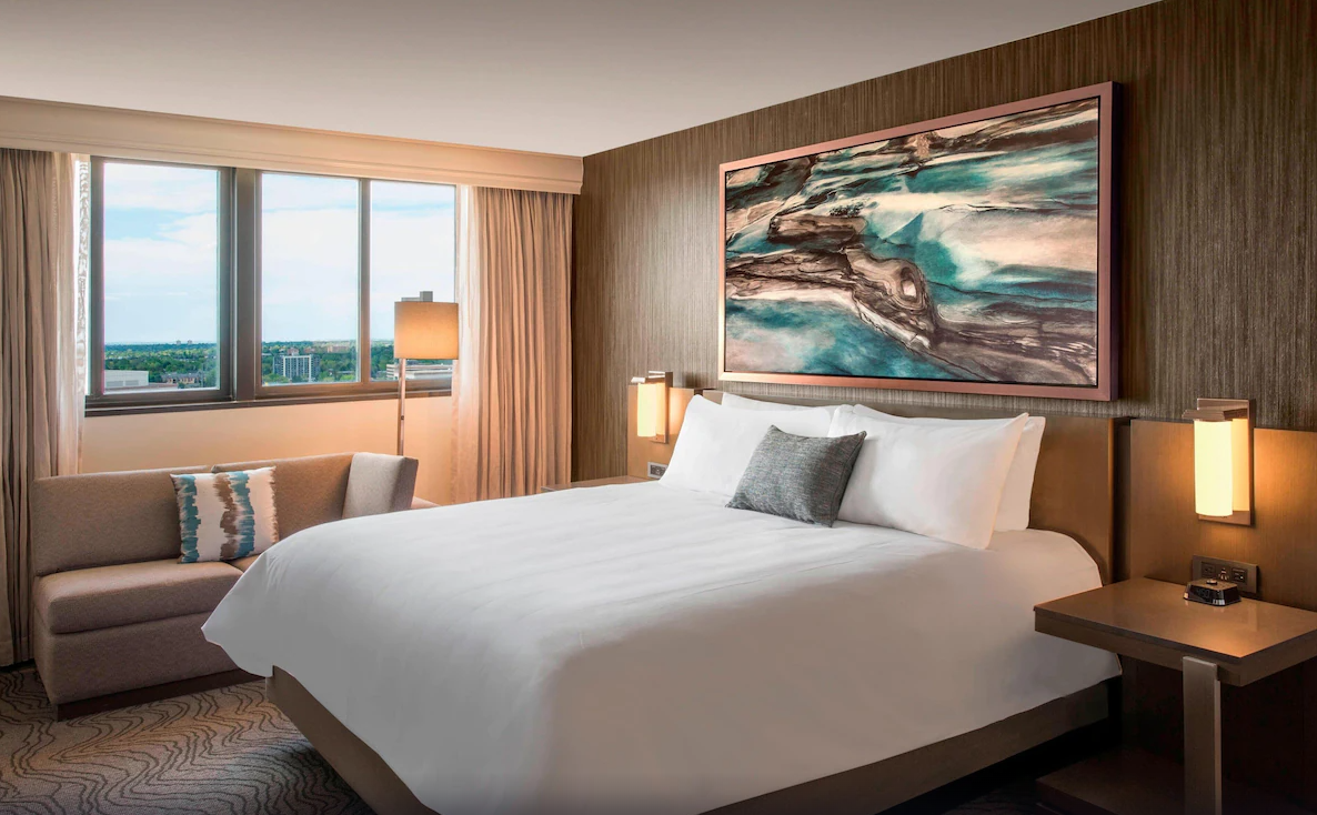 Modern hotel room with a large bed, abstract artwork, a sofa, and a view of Denver skyline through large windows.